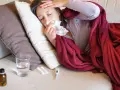 Kobieta z gorączką zas 53, chora, leży pod kocem, trzymając chusteczkę. Obok szklanka wody, termometr i leki.