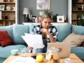 Kobieta w kratę siedzi na kanapie, analizując dokumenty przy laptopie.