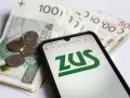 Telefon z logo ZUS na tle polskich banknotów i monet.