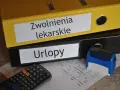 Dwa segregatory z napisami "Zwolnienia lekarskie" i "Urlopy", kalkulator, pieczątka i dokumenty.
