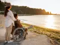 Kobieta w kapeluszu obejmuje ramieniem mężczyznę na wózku inwalidzkim, patrzą na zachód słońca nad morzem.
