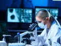 Kobieta w masce i okularach ochronnych pracuje przy mikroskopie w laboratorium.