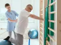 Fizjoterapeutka pomaga pacjentowi ćwiczyć na piłce rehabilitacyjnej przy drabinkach.