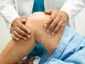 Fizjoterapeuta delikatnie masuje kolano pacjenta w niebieskiej koszuli.