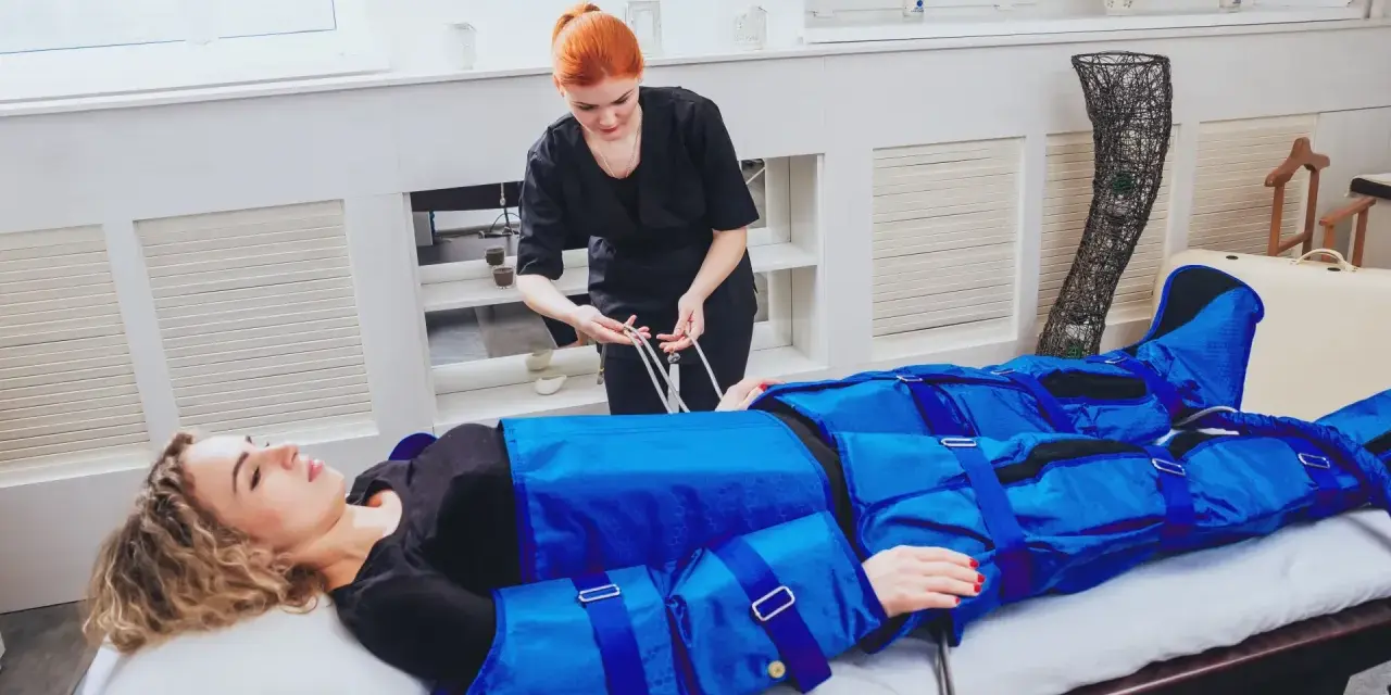 Kobieta w kombinezonie do drenażu limfatycznego leży na łóżku, a terapeutka podłącza przewody.