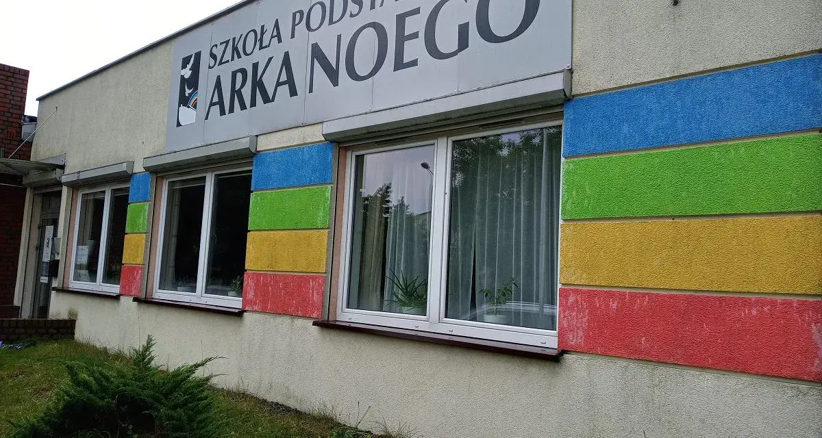 Budynek szkoły z kolorowymi pasami i napisem "SZKOŁA PODSTAWOWA ARKA NOEGO".