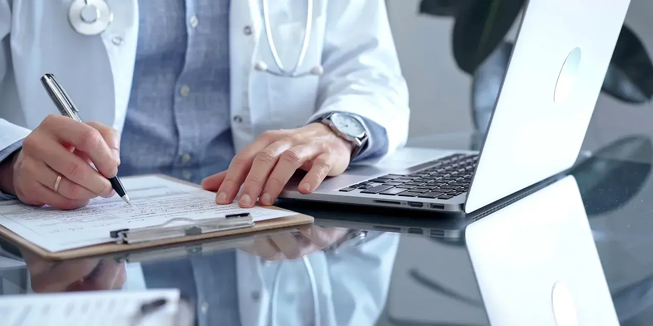 Lekarz w białym kitlu pisze na dokumentach, obok laptop. Zastanawia się, ile można być na zwolnieniu lekarskim od psychiatry.