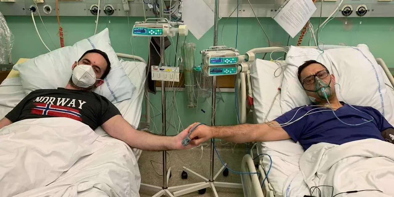 Dwóch mężczyzn w szpitalnych łóżkach, po przeszczepie nerki, trzyma się za ręce. Jeden ma maskę na twarzy, drugi tlenową.