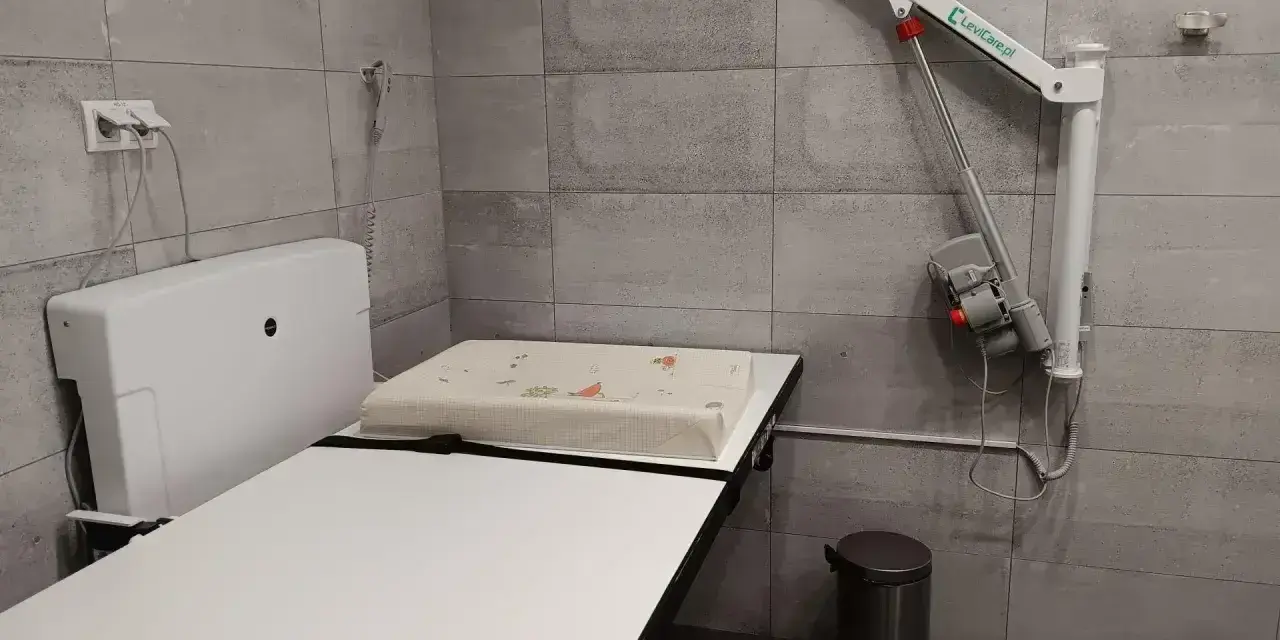 Przewijak dla dorosłych z podnośnikiem, koszem na śmieci i uchwytem na ścianie.