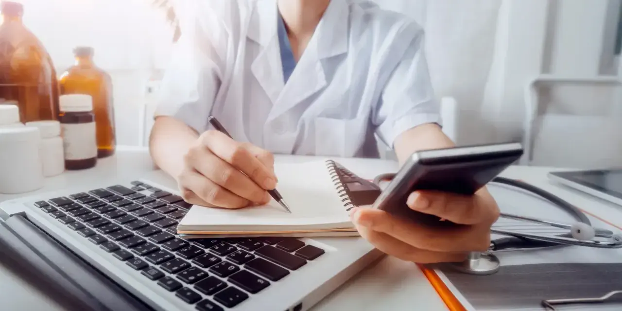 Lekarz w białym kitlu pisze w notatniku, trzymając telefon. Obok laptop, stetoskop i butelki.