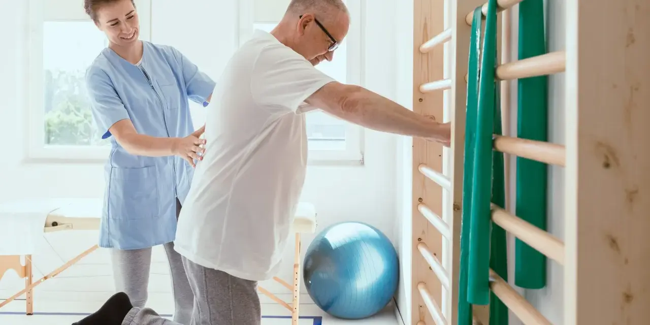 Fizjoterapeutka pomaga pacjentowi ćwiczyć na piłce rehabilitacyjnej przy drabinkach.