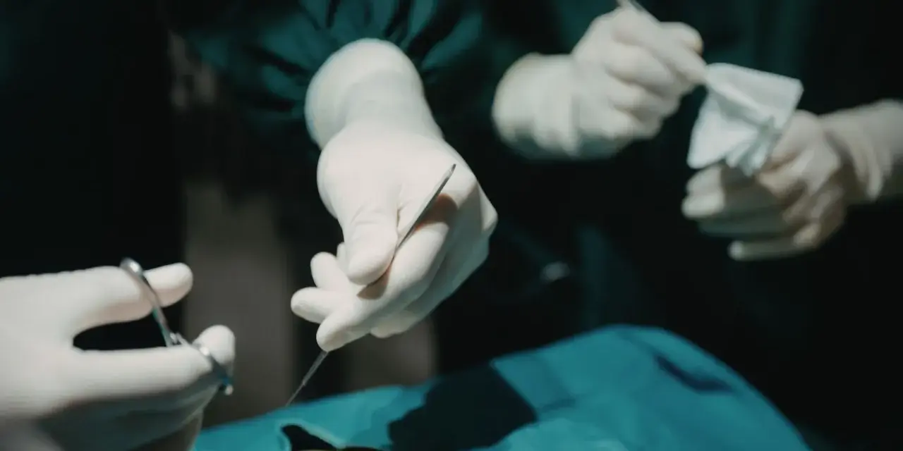 Dłonie chirurga w rękawiczkach trzymające narzędzia chirurgiczne i gazę podczas operacji.