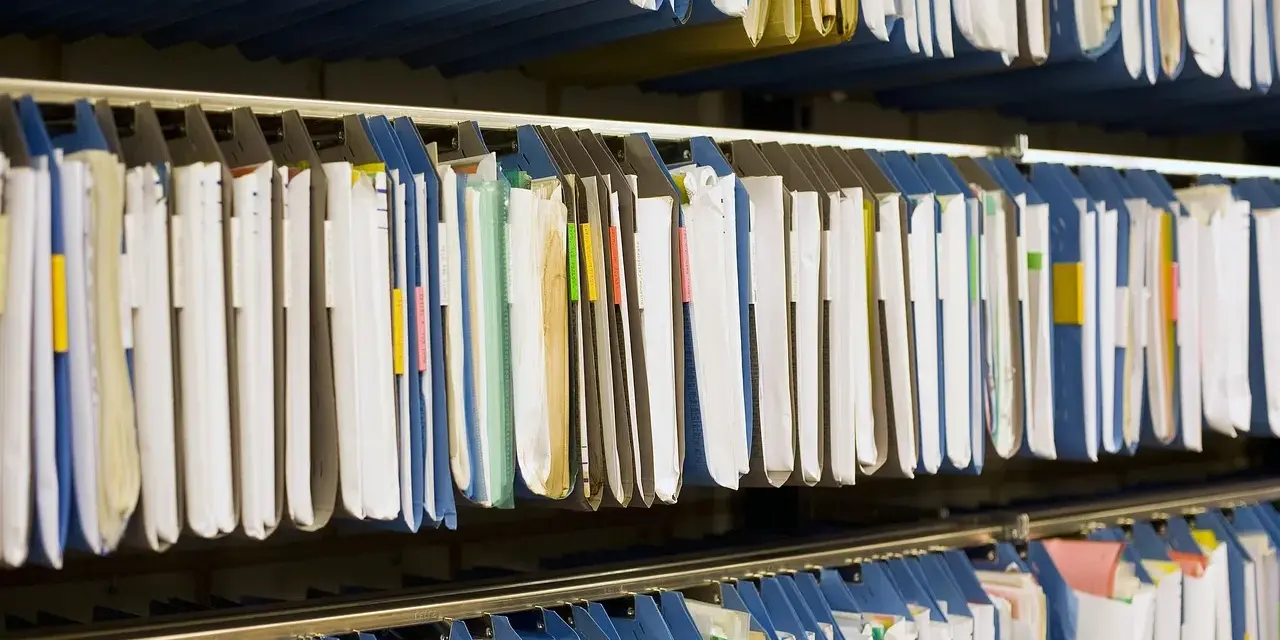 Rzędy wiszących segregatorów, gotowych do archiwizacji dokumentów. Widać tu wiele teczek, niektóre z kolorowymi etykietami, idealne do organizacji pracy, np. z wykorzystaniem systemu druk erp-6.