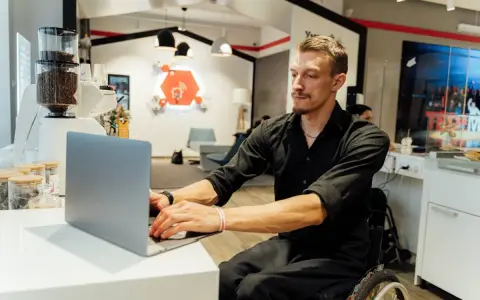 Mężczyzna na wózku inwalidzkim pracuje przy laptopie. To dowód, że praca z orzeczeniem o niepełnosprawności jest możliwa i satysfakcjonująca.