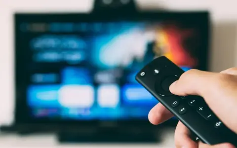 Dłoń trzyma pilota, celując w ekran telewizora. Szukasz sposobu, jak wyłączyć lektora dla niewidomych?