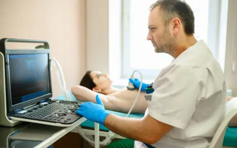 Lekarz wykonuje badanie USG piersi. Pacjentka leży na kozetce, a lekarz w rękawiczkach przykłada głowicę aparatu. Badanie jest ważne dla kobiet, które przeszły mastektomię, pomagając w diagnostyce i monitorowaniu stanu zdrowia, co może wpływać na ich g...