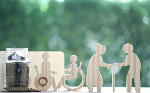 Skarbonka i drewniane figurki symbolizujące osoby w różnym wieku i z niepełnosprawnością. Czy świadczenie wspierające wlicza się do dochodu?
