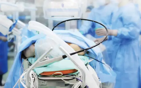 Pacjent na stole operacyjnym, przygotowany do zabiegu PCI kardiologia. W tle personel medyczny w niebieskich fartuchach.
