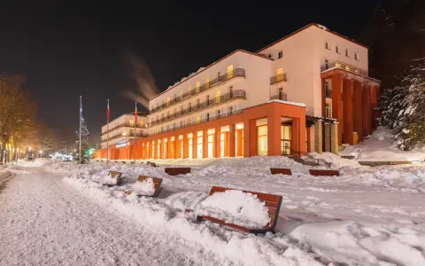 Budynek sanatorium nocą, ośnieżony teren z ławkami i drzewami.