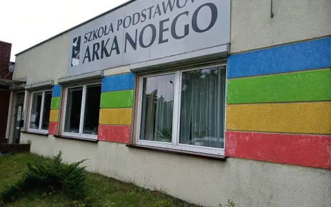 Budynek szkoły z kolorowymi pasami i napisem "SZKOŁA PODSTAWOWA ARKA NOEGO".