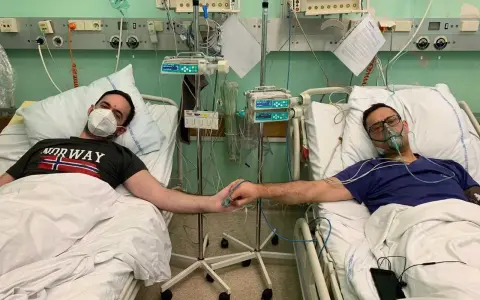 Dwóch mężczyzn w szpitalnych łóżkach, po przeszczepie nerki, trzyma się za ręce. Jeden ma maskę na twarzy, drugi tlenową.