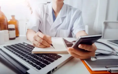 Lekarz w białym kitlu pisze w notatniku, trzymając telefon. Obok laptop, stetoskop i butelki.