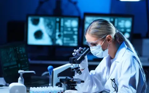 Kobieta w masce i okularach ochronnych pracuje przy mikroskopie w laboratorium.