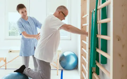 Fizjoterapeutka pomaga pacjentowi ćwiczyć na piłce rehabilitacyjnej przy drabinkach.