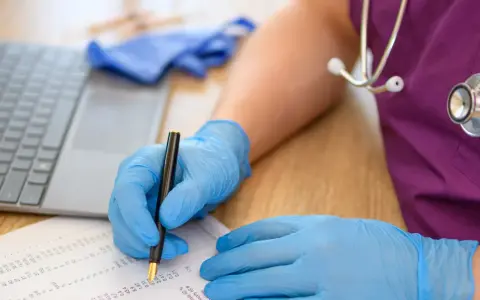 Dłoń w niebieskiej rękawiczce pisze piórem na dokumentach obok laptopa i stetoskopu.