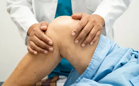Fizjoterapeuta delikatnie masuje kolano pacjenta w niebieskiej koszuli.