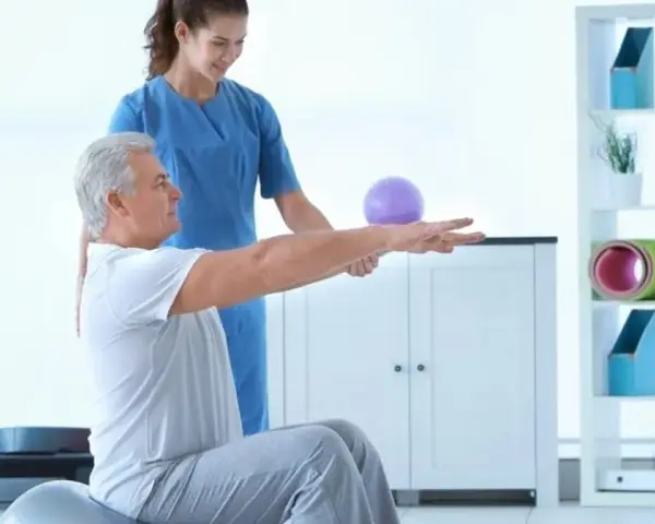 Fizjoterapeutka pomaga starszemu mężczyźnie ćwiczyć na piłce rehabilitacyjnej.