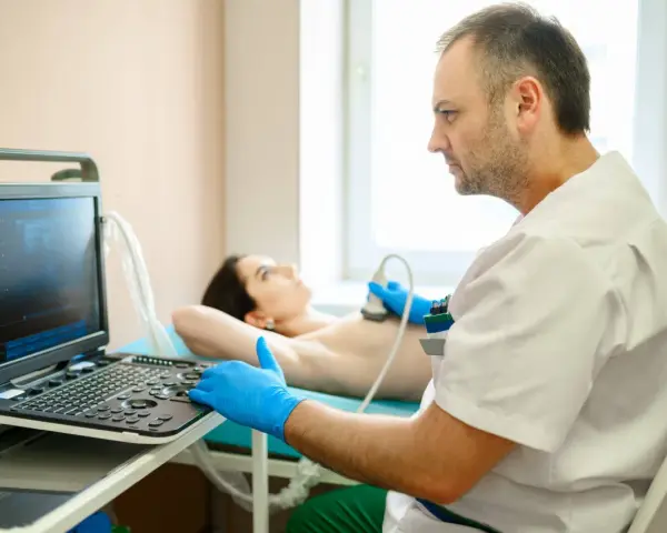 Lekarz wykonuje badanie USG piersi. Pacjentka leży na kozetce, a lekarz w rękawiczkach przykłada głowicę aparatu. Badanie jest ważne dla kobiet, które przeszły mastektomię, pomagając w diagnostyce i monitorowaniu stanu zdrowia, co może wpływać na ich g...