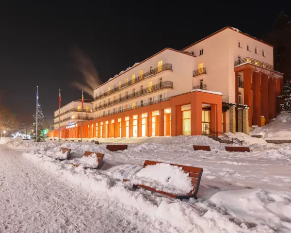 Budynek sanatorium nocą, ośnieżony teren z ławkami i drzewami.
