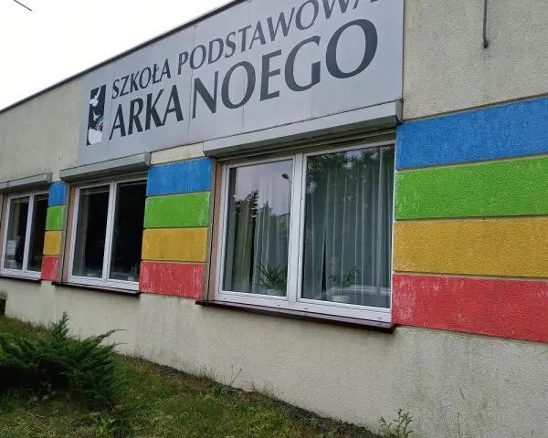 Budynek szkoły z kolorowymi pasami i napisem "SZKOŁA PODSTAWOWA ARKA NOEGO".