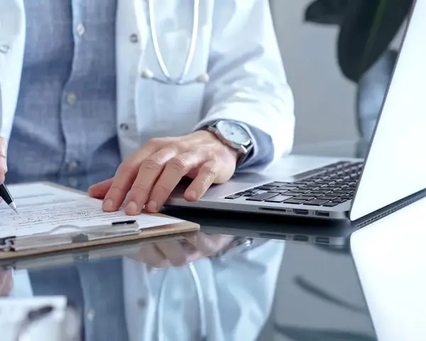 Lekarz w białym kitlu pisze na dokumentach, obok laptop. Zastanawia się, ile można być na zwolnieniu lekarskim od psychiatry.