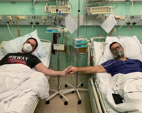 Dwóch mężczyzn w szpitalnych łóżkach, po przeszczepie nerki, trzyma się za ręce. Jeden ma maskę na twarzy, drugi tlenową.