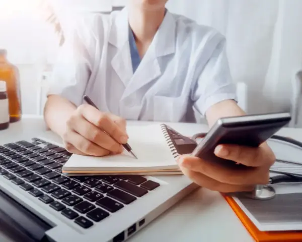 Lekarz w białym kitlu pisze w notatniku, trzymając telefon. Obok laptop, stetoskop i butelki.