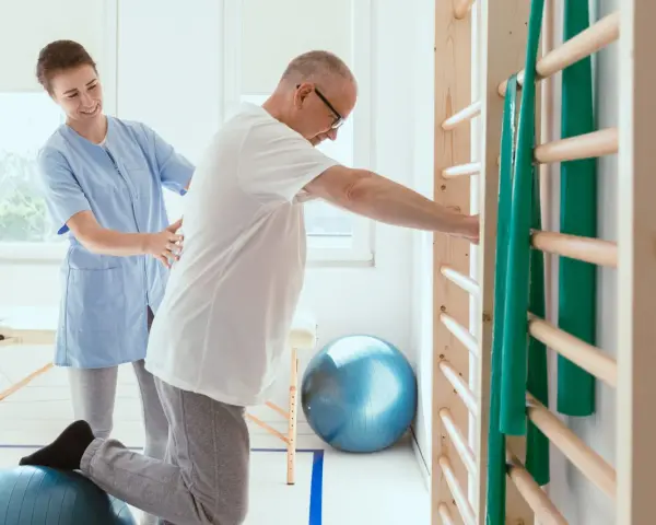 Fizjoterapeutka pomaga pacjentowi ćwiczyć na piłce rehabilitacyjnej przy drabinkach.