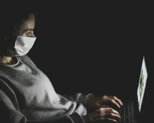 Kobieta w masce medycznej pracuje na laptopie w ciemności.
