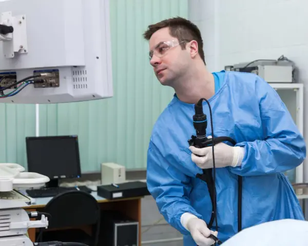 Lekarz w niebieskim fartuchu i okularach ochronnych trzyma endoskop, patrząc na monitor.