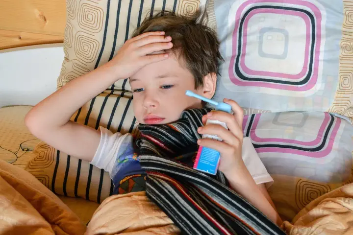 Chory chłopiec leży w łóżku z ręką na czole i trzyma inhalator.