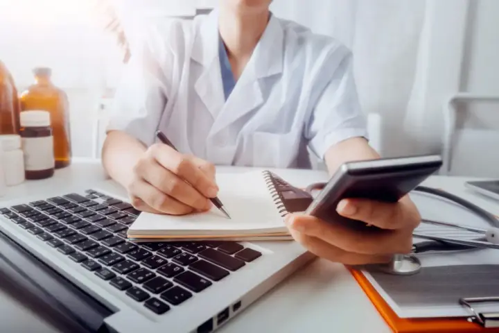 Lekarz w białym kitlu pisze w notatniku, trzymając telefon. Obok laptop, stetoskop i butelki.