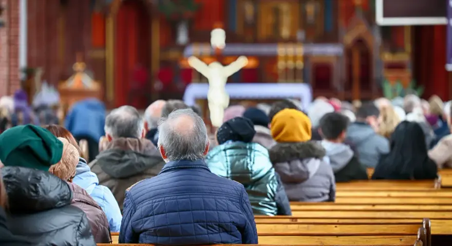 Ludzie siedzący w kościele, widok od tyłu, w tle ołtarz z krzyżem.