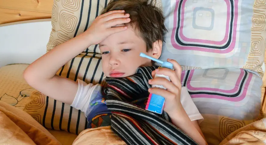 Chory chłopiec leży w łóżku z ręką na czole i trzyma inhalator.