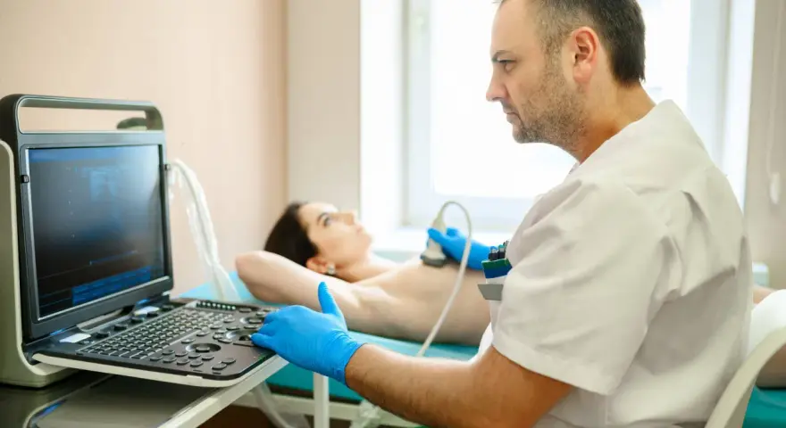 Lekarz wykonuje badanie USG piersi. Pacjentka leży na kozetce, a lekarz w rękawiczkach przykłada głowicę aparatu. Badanie jest ważne dla kobiet, które przeszły mastektomię, pomagając w diagnostyce i monitorowaniu stanu zdrowia, co może wpływać na ich g...