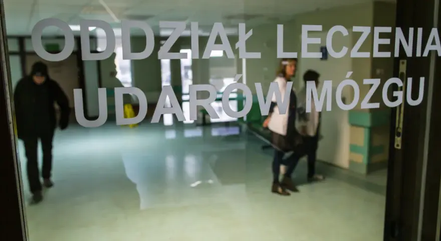 Drzwi do oddziału leczenia udarów mózgu. W środku widać personel medyczny i pacjentów, gotowych na zabiegi takie jak trombektomia.