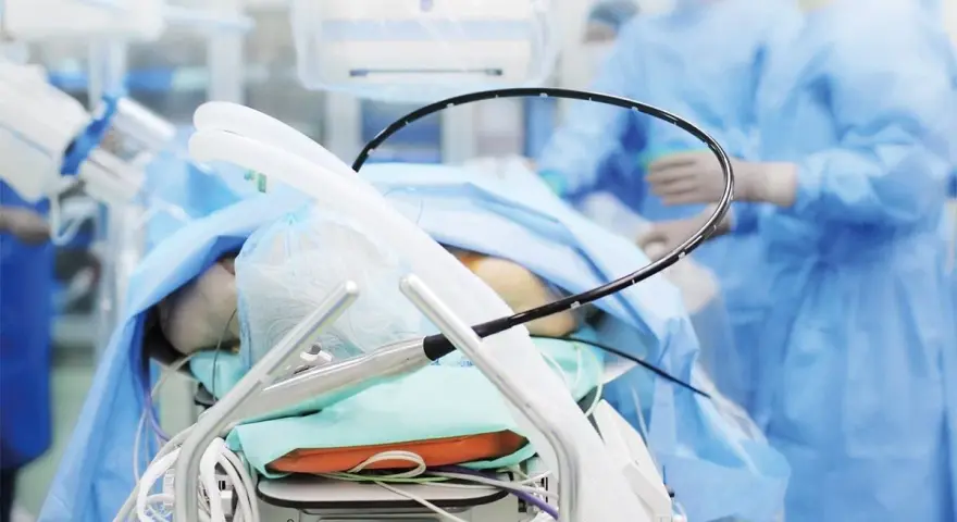 Pacjent na stole operacyjnym, przygotowany do zabiegu PCI kardiologia. W tle personel medyczny w niebieskich fartuchach.