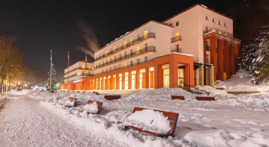 Budynek sanatorium nocą, ośnieżony teren z ławkami i drzewami.