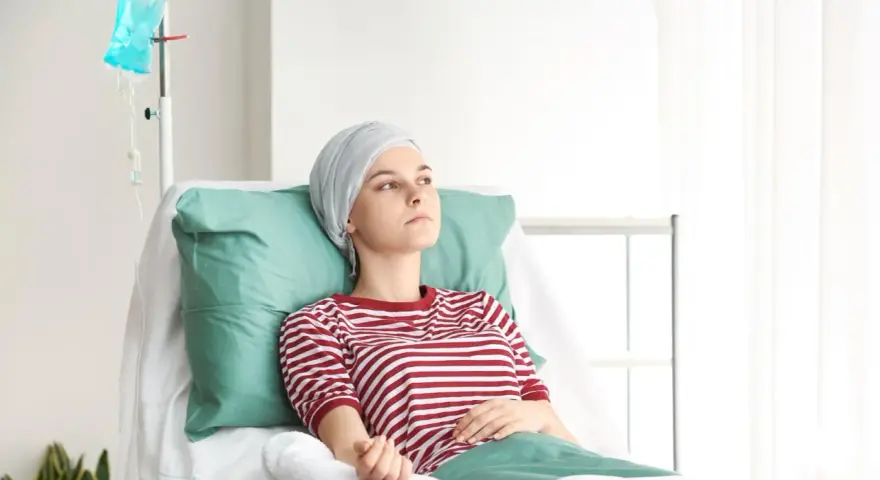Młoda kobieta w chuście na głowie leży w szpitalnym łóżku, podłączona do kroplówki.