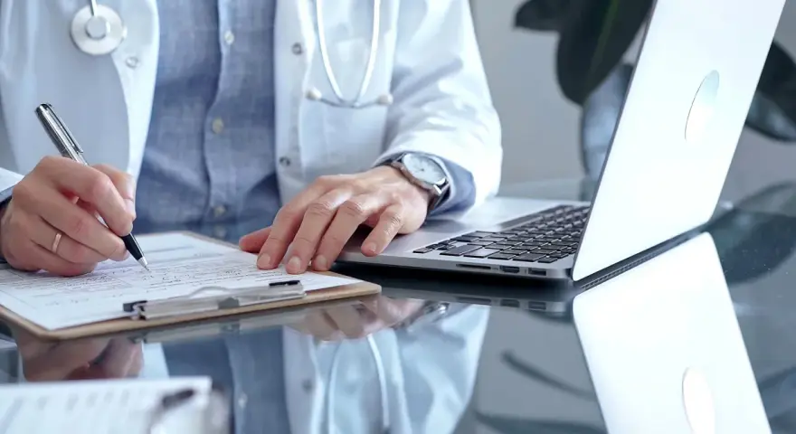 Lekarz w białym kitlu pisze na dokumentach, obok laptop. Zastanawia się, ile można być na zwolnieniu lekarskim od psychiatry.